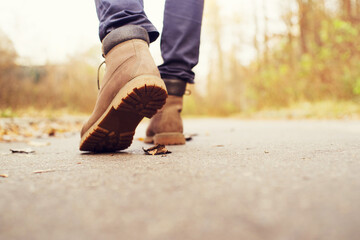Feet in brown leather boots are walking along the road on an autumn day, close up, back view, soft focus