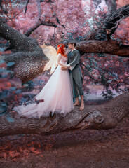Young couple in love. A man and a woman are hugging on a huge tree. Themed creative wedding bright fantasy photography. Fairy woman in long pink dress with bright golden wings. A man in an elf costume © kharchenkoirina