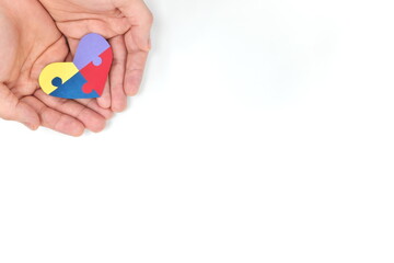Cupped male hands holding a colorful puzzle heart shape paper cutout. Top view flat lay in white background with copy space. World Autism awareness day, support and care concept.