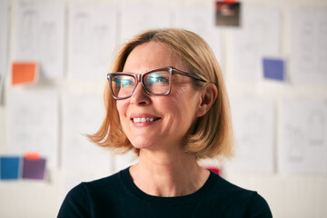 Portrait Of Mature Woman Standing In Front Of Designs On Wall In Start Up Fashion Business