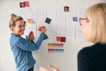 Female Business Owner With Intern Looking At Designs On Wall In Start Up Fashion Business