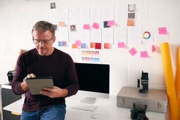 Mature Male Photographer Using Digital Tablet Sitting On Stool In Front Of Desk In Studio