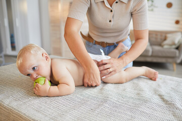 Unrecognizable modern mother changing diapers of her little baby son while he is biting green apple