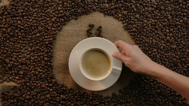 Symbol of hot coffee cup drawn with roasted arabica grains turns into real cup with strong espresso by waiter or barista hand. Creative coffee transition. Flat lay view. Slow motion.