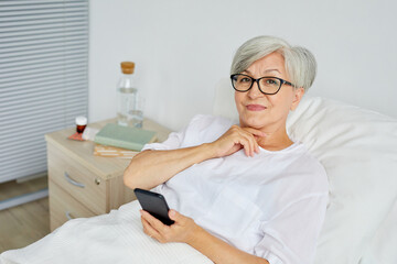 Fototapeta premium Portrait of beautiful aged woman wearing white clothes lying on bed in hospital ward holding smartphone looking at camera