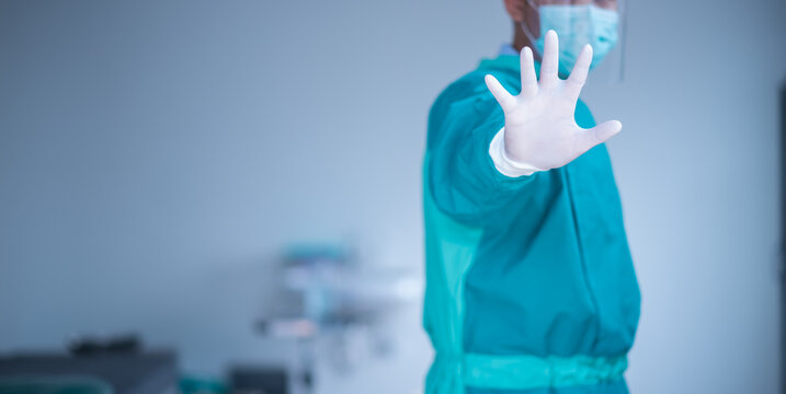 male doctor in a coat and  sterile gloves shows a stop gesture with his right hand, white background