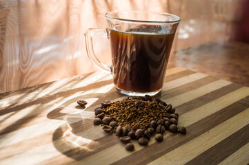 Brewed black coffee stands in a transparent cup on a wooden plank with whole, roasted beans and instant coffee scattered on it.