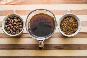 There are three cups of coffee on a wooden board, one with roasted black coffee beans, the second with brewed coffee, and the third with instant coffee.
