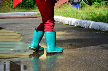 little funny cute girl walks in the rain with a red umbrella, in green rubber boots through the puddles. Laughs, has fun. girl wearing yellow waterproof coat playing on a warm autumn or summer day