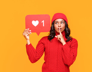 Young ethnic female showing banner with like symbol and keeping finger near lips while sharing secret in social media against yellow background 