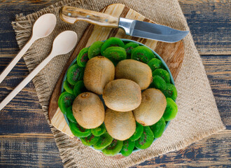 Kiwi with dried slices, cutting board, knife, spoons in a plate on wooden and piece of sack background, flat lay.