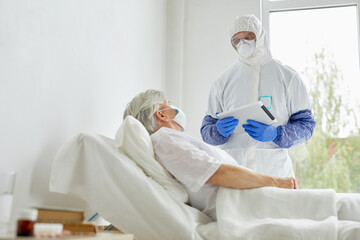 Unrecognizable man wearing protective suit with gloves, mask and eyewear holding digital tablet working with patient in infectious disease ward