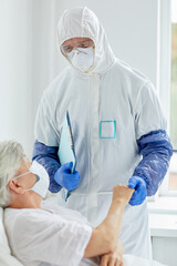 Fototapeta premium Professional doctor wearing protective suit with gloves examining health of senior woman lying on bed in infectious disease ward
