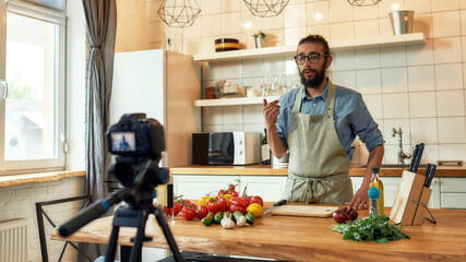 Young man, Italian cook in apron looking at camera, filming himself for culinary blog while preparing healthy meal with vegetables in the kitchen