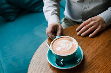 Fototapeta na wymiar Hands of a businessman freelancer with cup of coffee on a table. Blue cup of Coffee with foam and picture