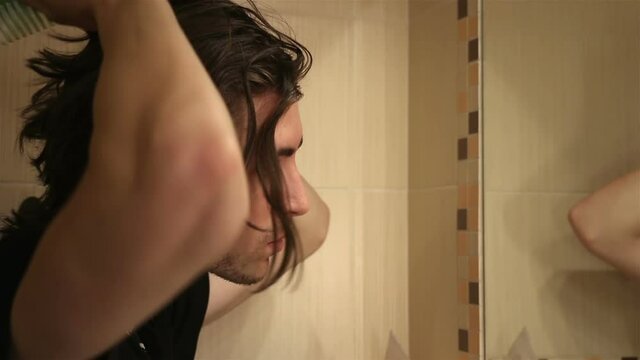 Handsome young man brushing long layered dark brown hair in bathroom, standing in front of mirror, face profile