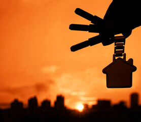 Buying or renting resindental house concept. Man hand holding 3 three silver key with houme shape keyring, on sunlight sky cityscapes background.