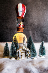 still life miniature cabin in snow surrounded by pine trees  and full moon in background with Santa in hot air balloon overhead