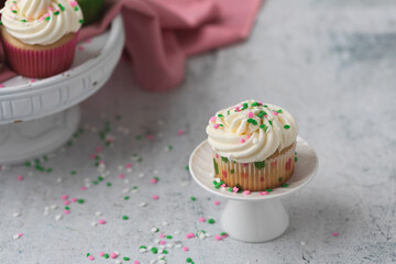 Vanilla Cupcake with White Icing and Sprinkles on a Small White Pedestal with Vanilla and Chocolate Cupcakes on a Stand in the Background