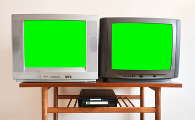 Two antiquated vintage TVs with VCRs with blank green screens sit on a vintage table in a 1990s tenement building.