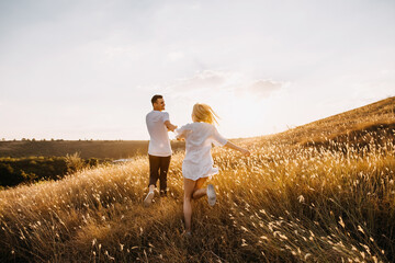 Young romantic couple running in an open field with dry grass at sunset, holding hands.