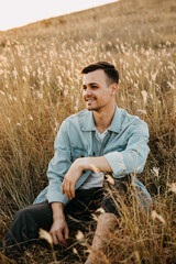 Naklejka na ściany i meble Young handsome man, wearing denim jacket, sitting in a field, looking to side, smiling.