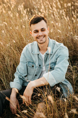 Naklejka na ściany i meble Young handsome man, wearing denim jacket, sitting in a field, smiling.