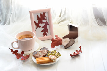 Coffee cap, chocolate chip cookies on a plate,wooden hearts,gift box,red autumn leaves on bage table by the window with transparent organza curtain