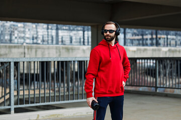 Attractive runner in shape in red tracksuit standing, listening music and holding refreshment. He is taking a break from running in the urban part of the city.