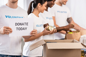 Selective focus of man holding card with donate today lettering near multiethnic volunteers and...