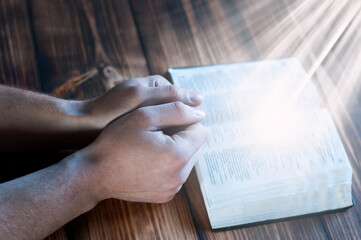 Naklejka premium Open holy bible on a wooden table. Hands in prayer. Light on the book