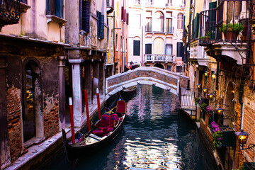 Obraz na płótnie Canvas Beautiful Venice canal with gondola and red brick bridge. The luxurious gondola has red velvet-lined seats. On one wall there are lit lights and purple flowers in the balconies.