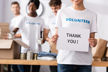 Selective focus of volunteer holding card with thank you lettering near donations and multiethnic...
