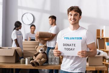 Obraz premium Selective focus of volunteer with donate today lettering on card looking at camera near multiethnic people in charity center