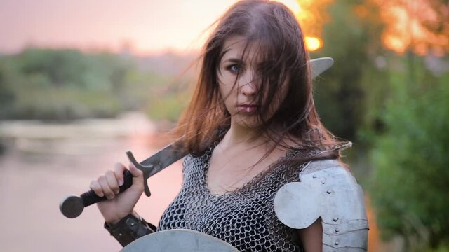 Camera movement from bottom to top, medieval woman warrior in chain mail armor with a shield and a sword in her hands posing while standing against the backdrop of bokeh sunset, river and forest.