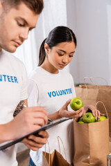 Selective focus of asian volunteer holding apples near man writing on clipboard in charity center