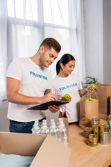Selective focus of multiethnic volunteers writing on clipboard and holding apples near tin cans and bottles of water on table
