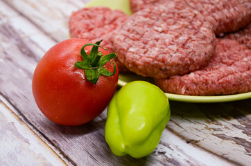 Ripe tomato and bell peppers on a background of raw homemade chopped beef burgers on a plate and a wooden table. BBQ preparation, beef products, traditional meat dish. Balanced diet. 