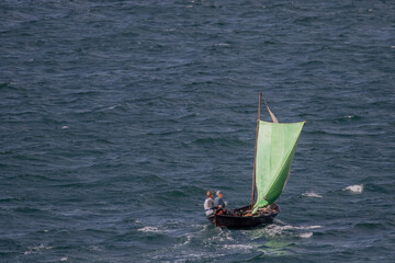 Two persons in a small wooden sailing boat in rough sea.