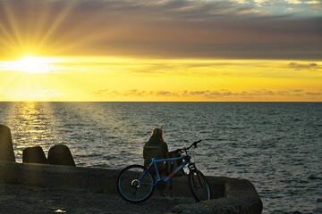 The girl is sitting on the edge of the pier and looks at the setting sun.