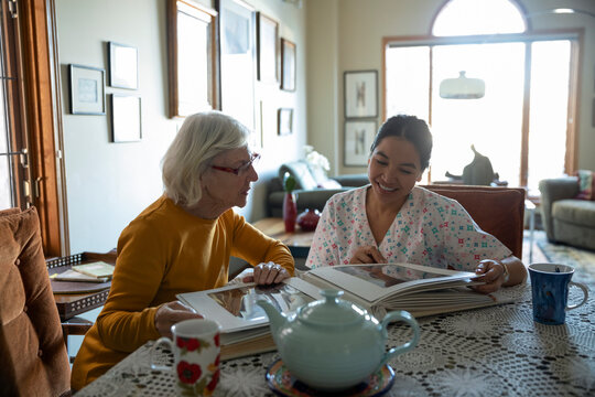 Home caregiver and senior woman looking at photo album at table