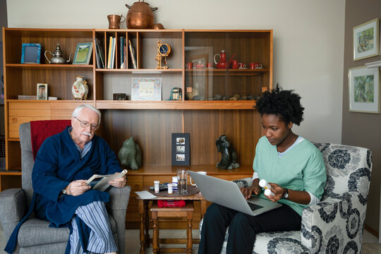 Home caregiver with laptop and senior man with magazine in living room