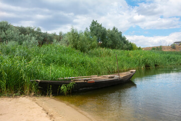 Fototapeta na wymiar Old wooden fisherman boat on the river. Beautiful summer landscape with high reeds on the shore, lake and sky with clouds. Retro ship on the water