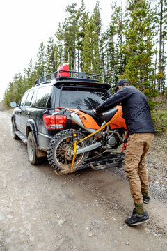 Man fastening dirt bike to overland SUV on dirt road in woods