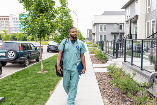 Male home caregiver in scrubs walking on sidewalk outside house
