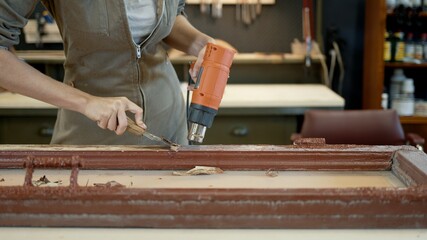 Restoration of wooden doors, window. A working female in worksuit carpenter peels off paint from a wooden product with a spatula and a heat gun.