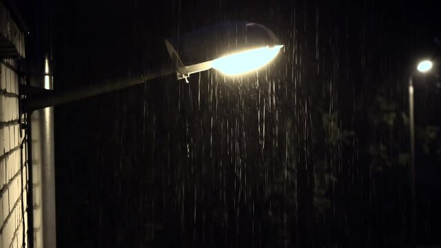 A street lamp attached to the wall of the house illuminates the road at night in the rain.