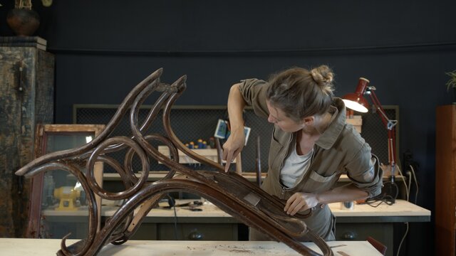 Restoration of wooden rocking chair in a workshop. A working female carpenter peels off paint from rocking chair legs with a spatula, restorer in a workshop