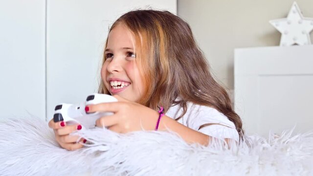 A little girl is playing a computer console on a sofa in a bright room. Concurrent games for children. Joystick for control in hands. Emotions and fun of the child. Copy space.High quality 4k footage.
