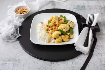 A Vegan Stir-Fry with Cauliflower and Peas on a White Plate, Black Tray, Cement Table, topped with Peanuts